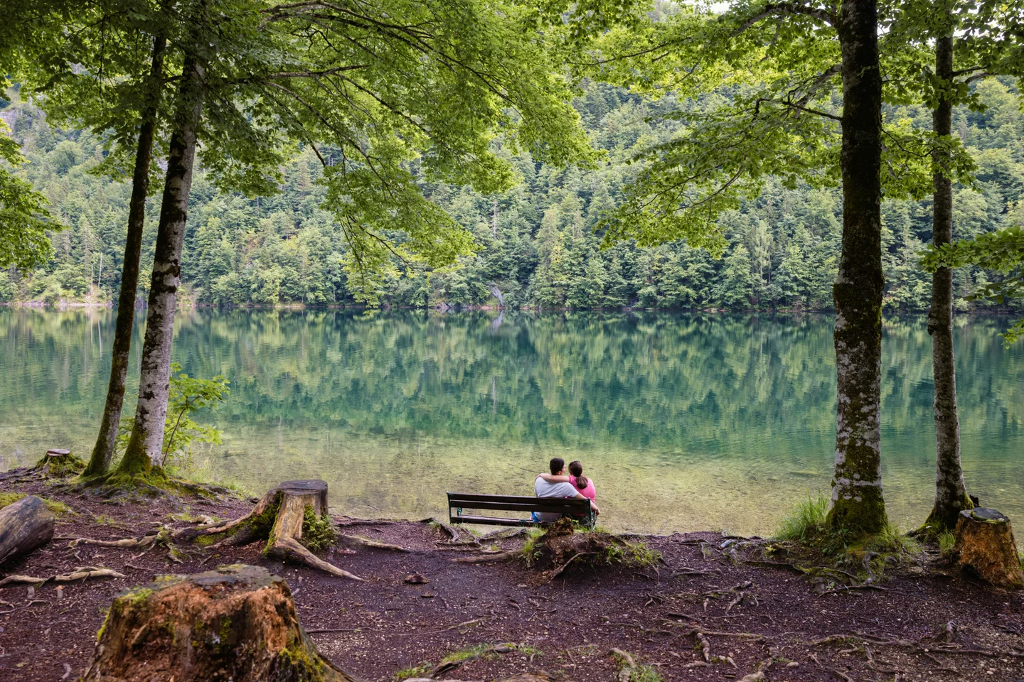 A couple sit with their arms around each other on a bench beside a calm, forest-lined lake.