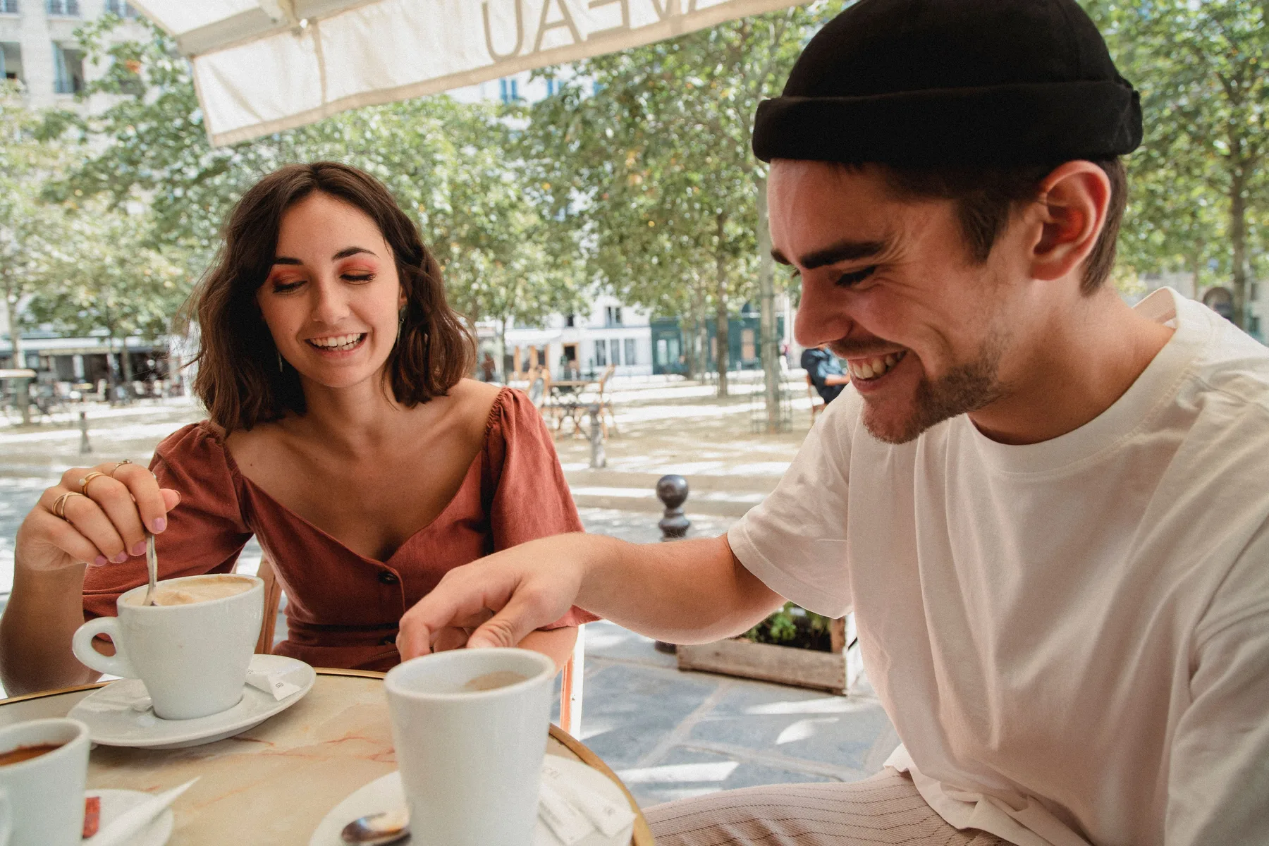 A smiling man and woman sit at a table over coffee in a sunny city café