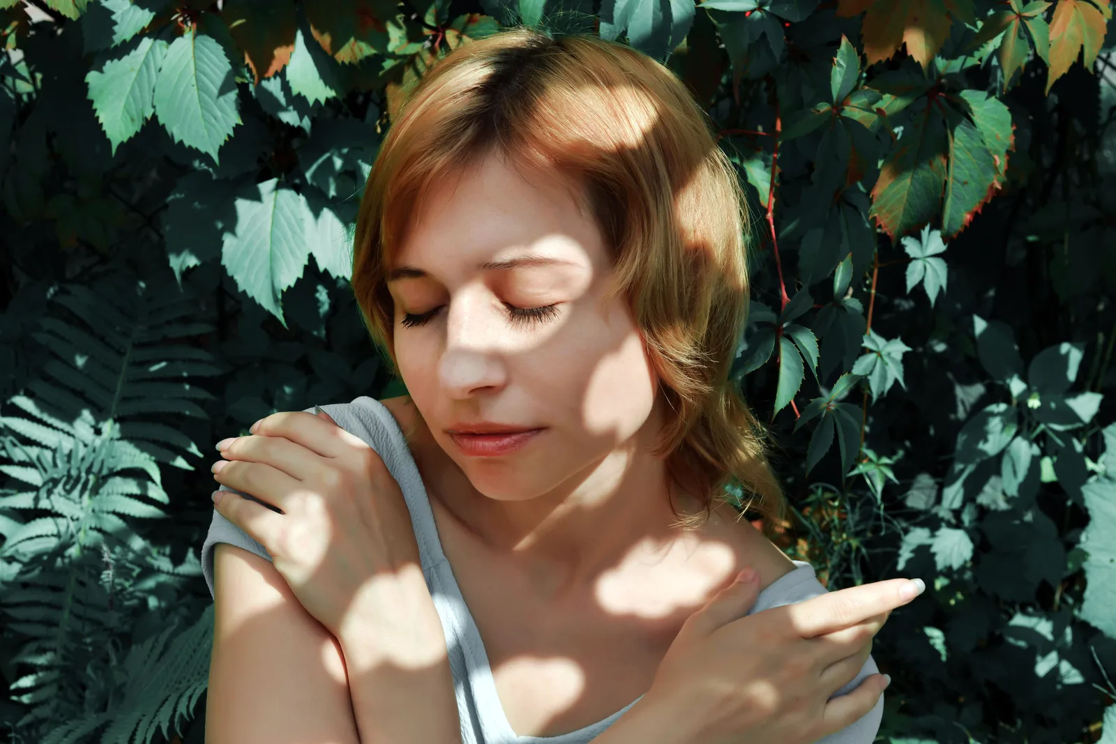 A woman with closed eyes rests her crossed hands on her shoulders in a sunlit green garden