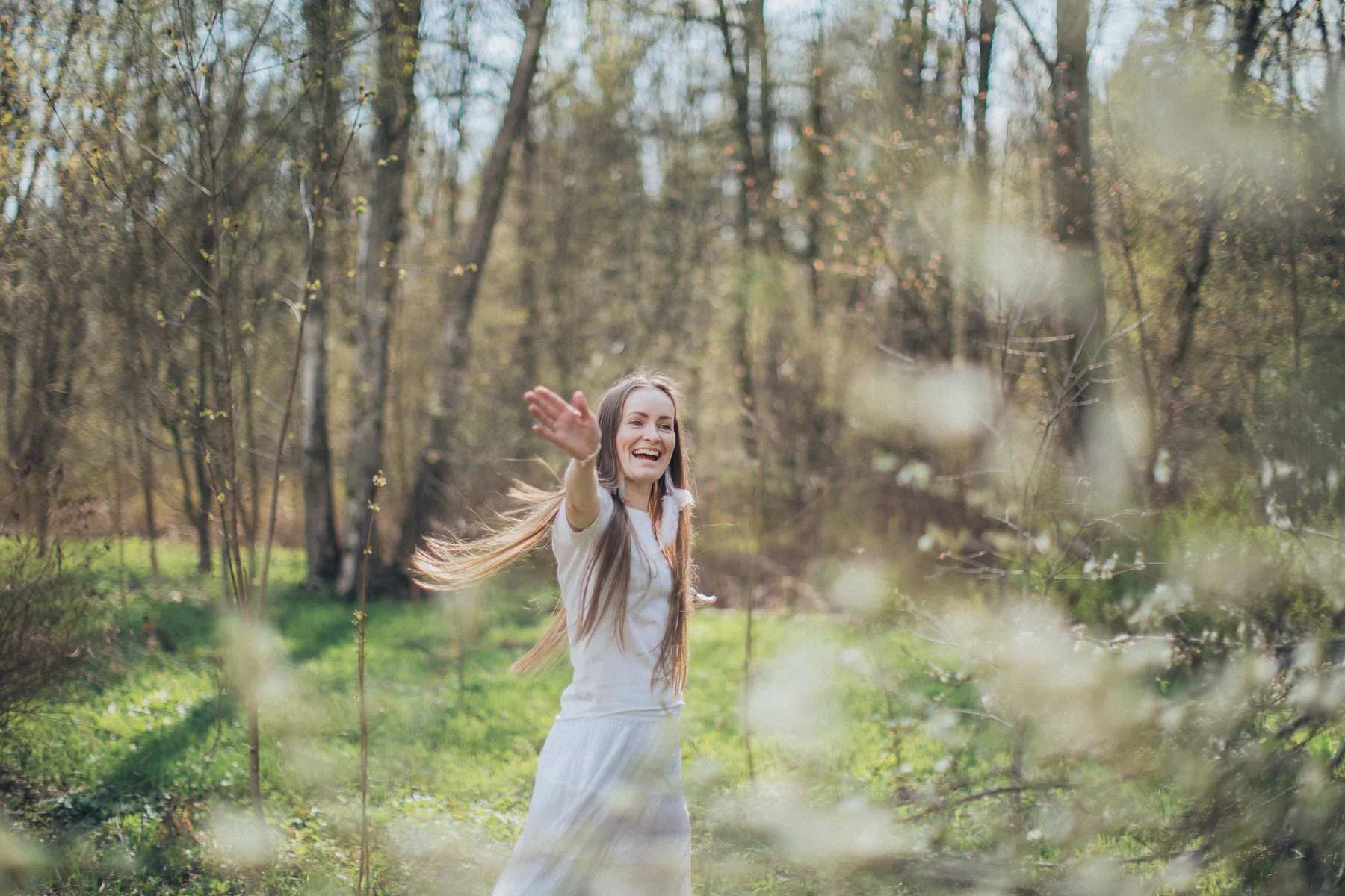 A smiling woman in white reaches out her hand as she turns in a sunlit forest