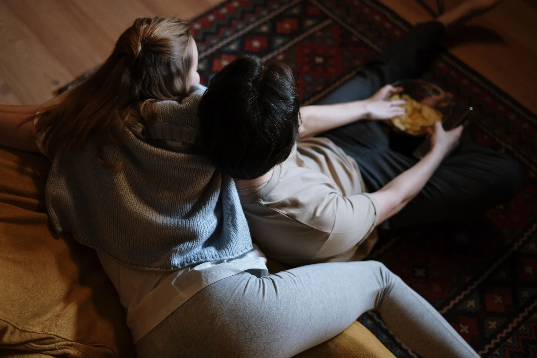 A couple sit cuddled on a sofa watching television with a bowl of snacks
